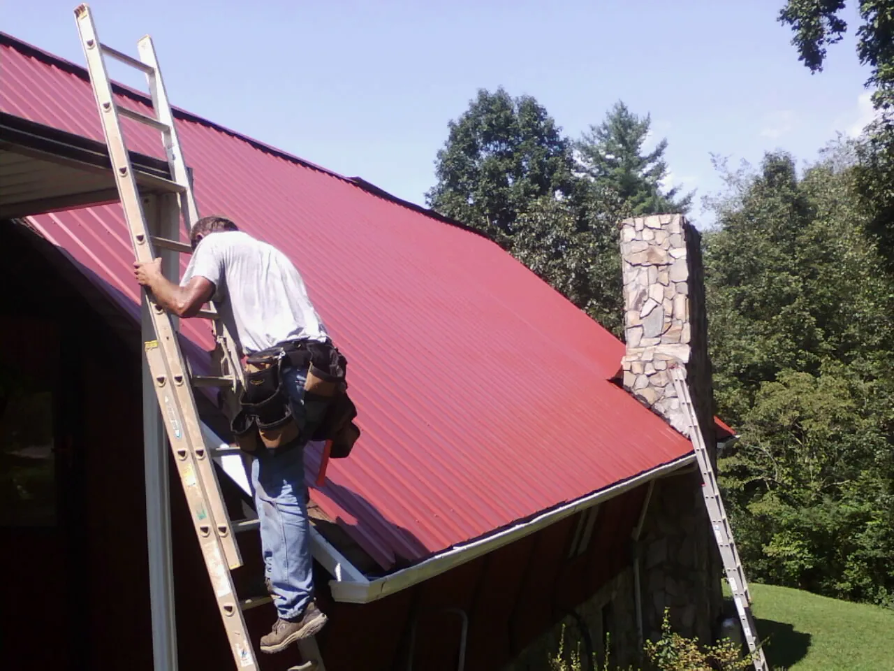 Roofer on a ladder beside a red metal roof and stone chimney — Soffit & Fascia Repair by Roofing Brothers in Urbanna
